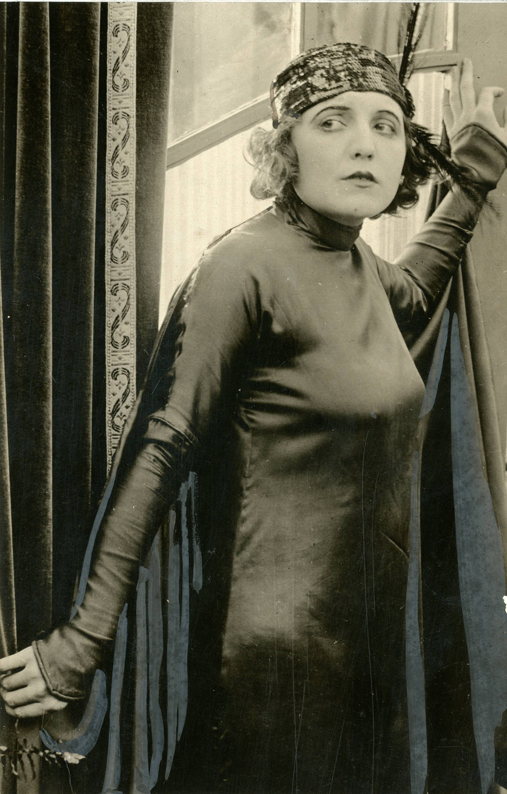 Black and white photo of a woman in a shiny, long-sleeved dress and a feathered headband, standing by a window with patterned curtains, looking to her left with a cautious expression.