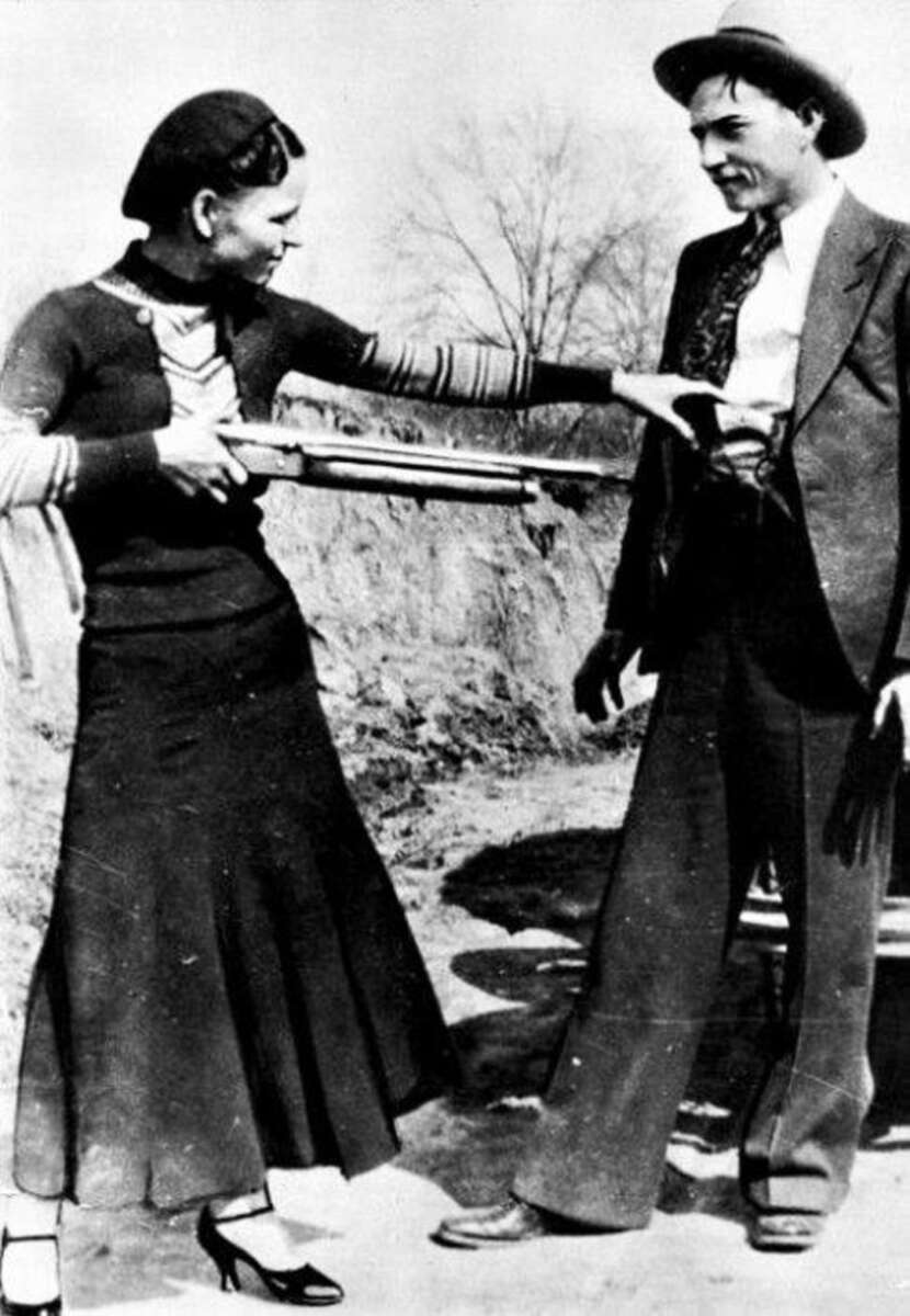A woman in a long skirt and heels playfully points a shotgun at a smiling man in a suit and hat outdoors; both appear relaxed and engaged with each other.