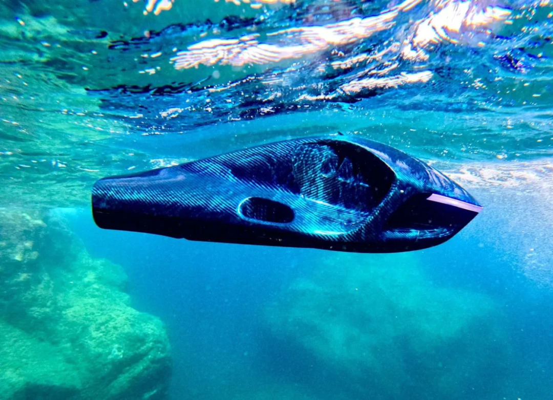 A streamlined, black underwater drone glides through clear turquoise water, with sunlight filtering from above and rocky seabed visible below.