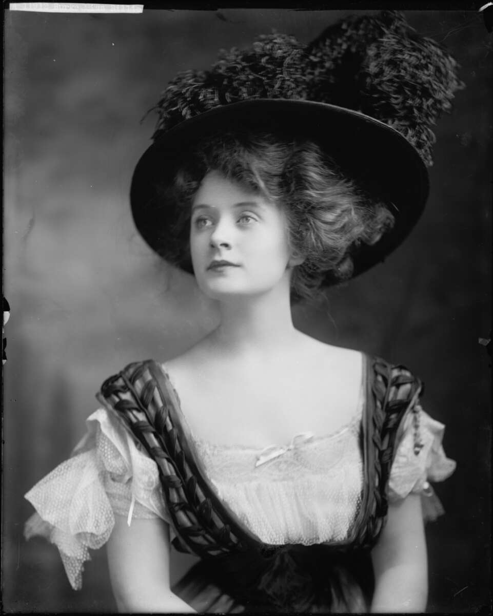 Black and white portrait of a young woman wearing a large, decorative hat with feathers and a dress with puffed sleeves and woven details. She gazes slightly upward with a calm, composed expression.