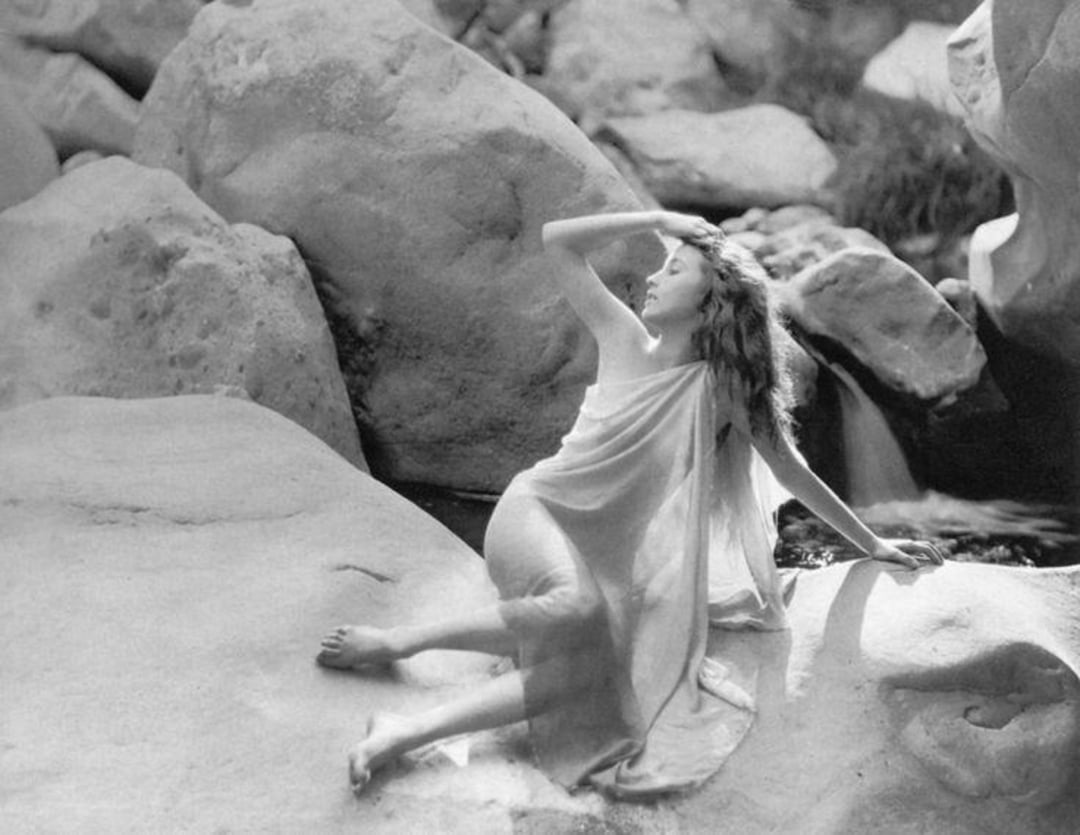 A woman draped in a light, flowing cloth sits on rocks by a stream, gazing upward with one arm shielding her eyes. Large, smooth boulders surround her in a natural, outdoor setting. The image is in black and white.