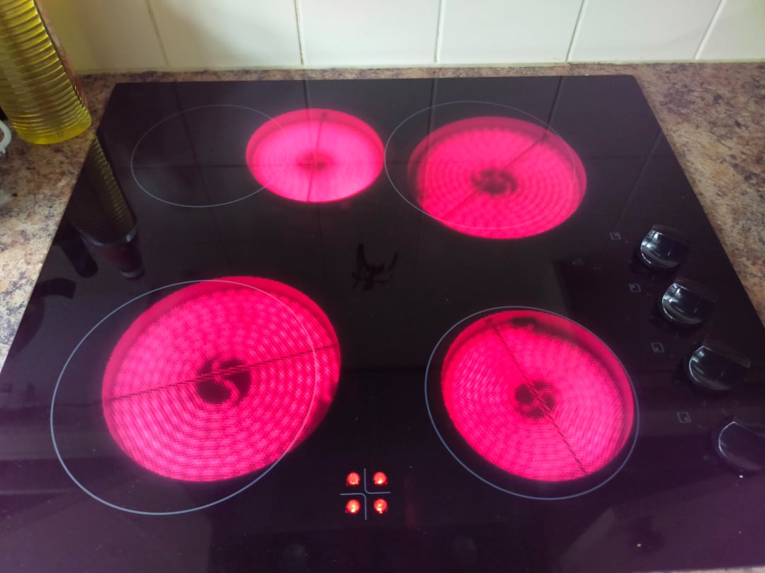 A black glass electric stovetop with four burners, all glowing red to indicate they are turned on and hot, set in a brown speckled countertop with white tiles in the background.
