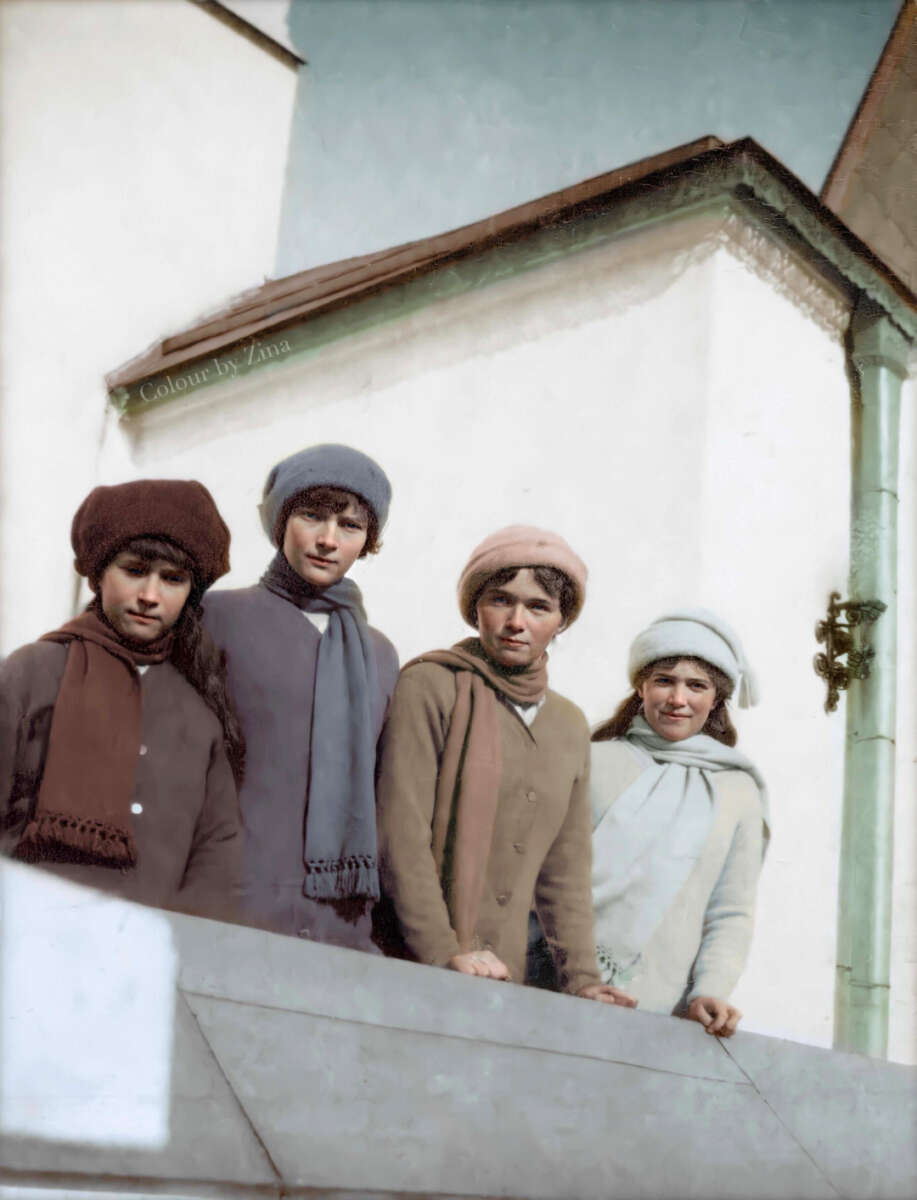 Four children in winter clothing—coats, scarves, and hats—stand side by side outdoors in front of a light-colored building, looking toward the camera. The image is colorized and has a vintage appearance.