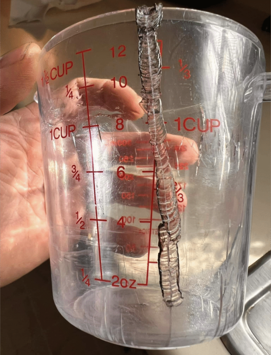 A clear plastic measuring cup with red markings is shown cracked and melted along one side, held up by a person’s hand. Light shines through the damaged area highlighting the melt pattern.