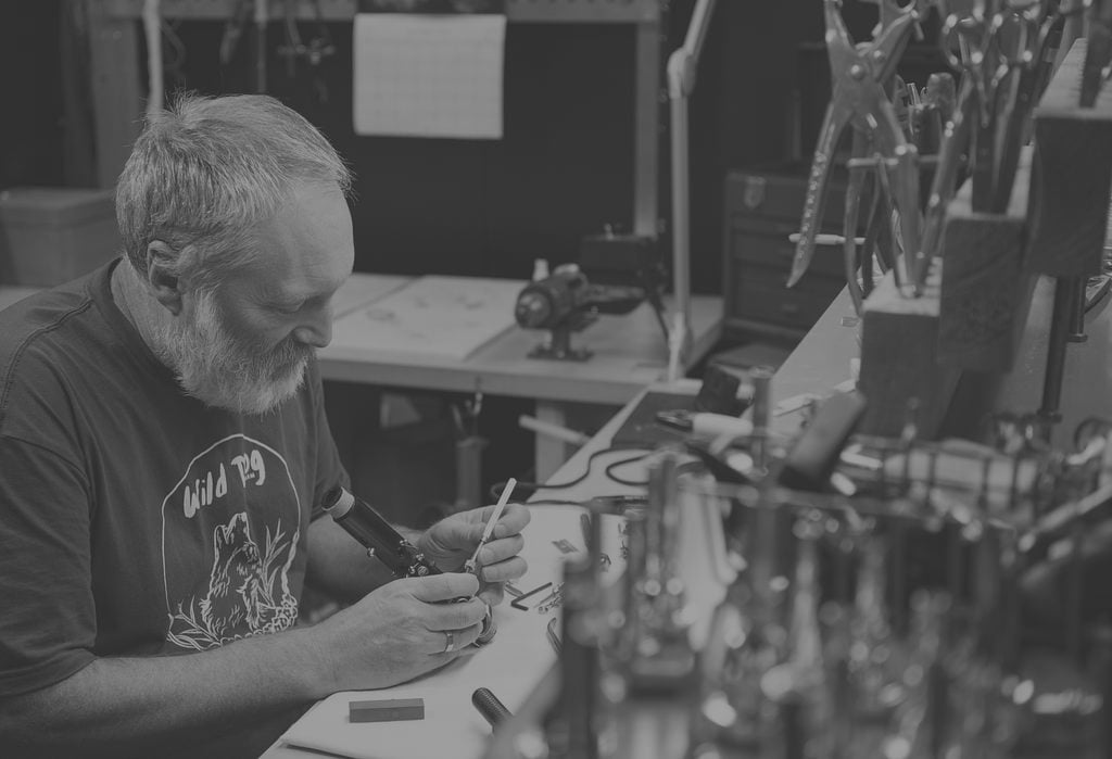 A bearded man sits at a cluttered workbench, intently working with tools in his hands. Various tools and equipment are visible around him, indicating a workshop or studio setting.