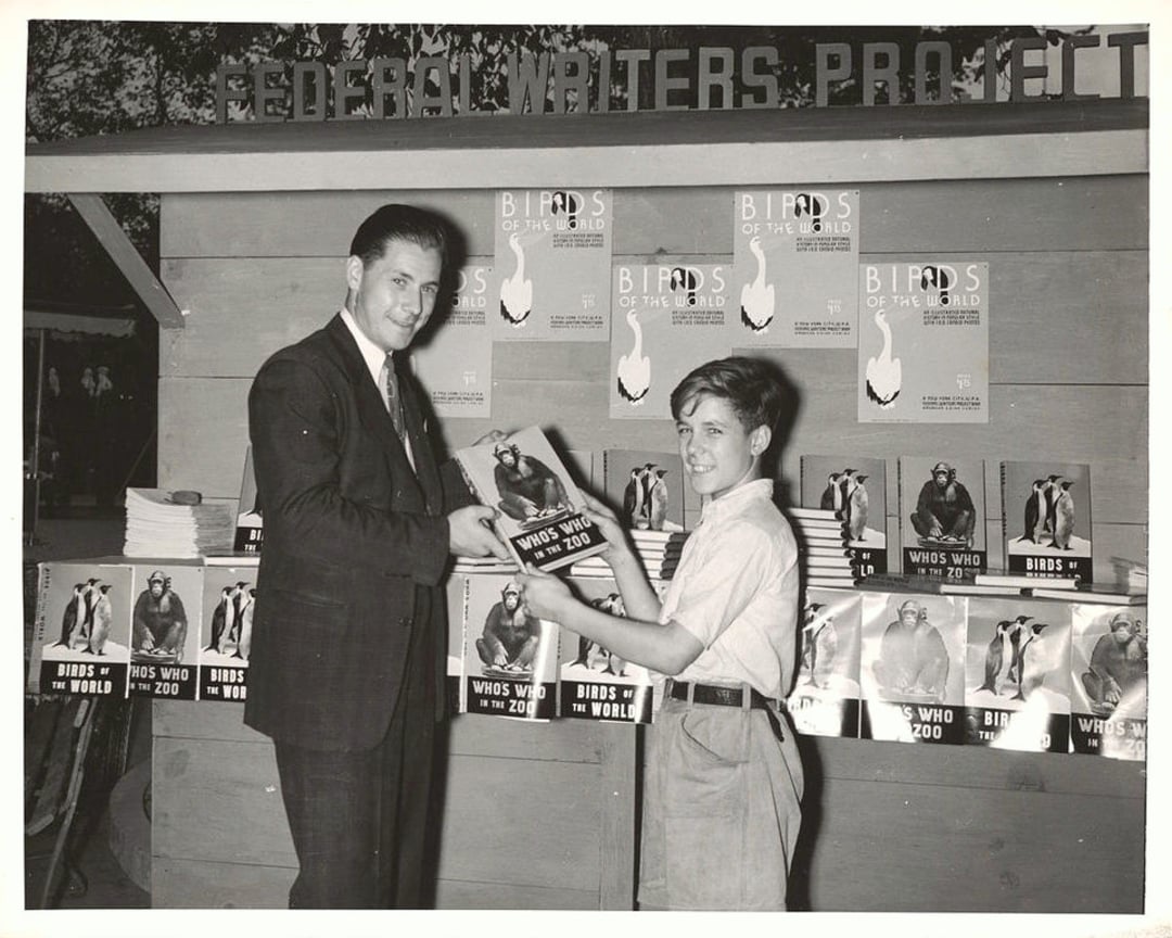 A man and a boy stand in front of a booth labeled "Federal Writers Project," smiling as the man hands the boy a book. The booth displays stacks of books and posters titled "Birds of the World" and "Who's Who in the Zoo.