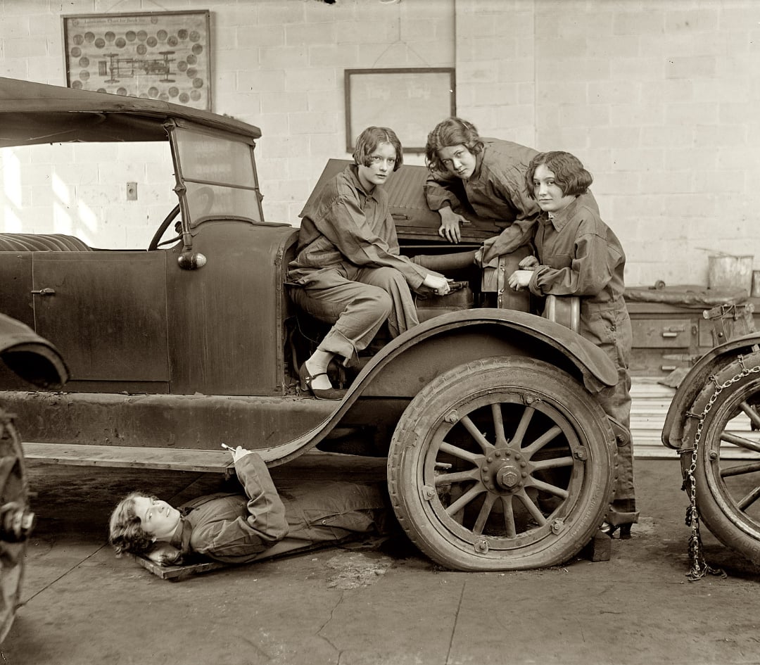 Four women in coveralls repair an old car in a garage; three are working around the hood, while one lies beneath the car. The scene appears to be from the early 20th century.