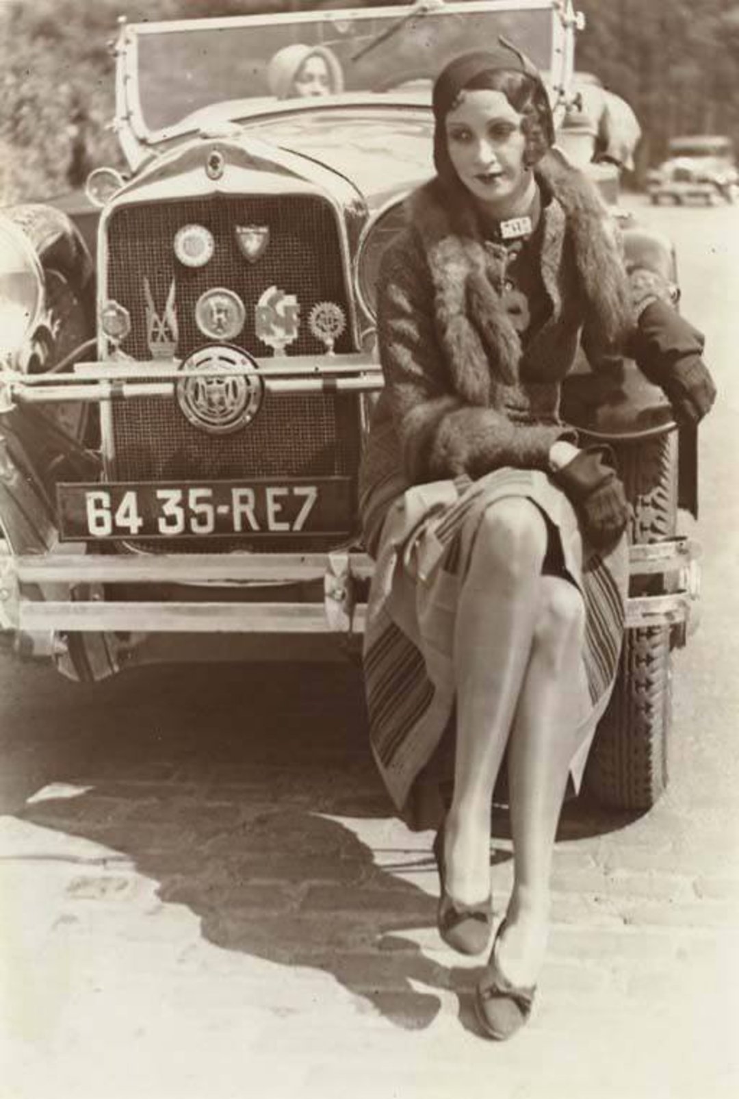 A stylish woman in 1920s fashion, wearing a fur-collared coat, cloche hat, and gloves, sits on the front bumper of a vintage car with a visible French license plate; another woman is seated inside the car.