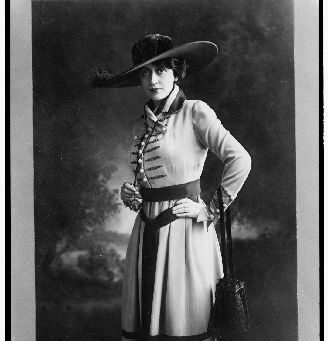 Black and white portrait of a woman standing, wearing a large brimmed hat and a long-sleeved dress with decorative buttons and a belt, posing with one hand on her hip against a painted backdrop.