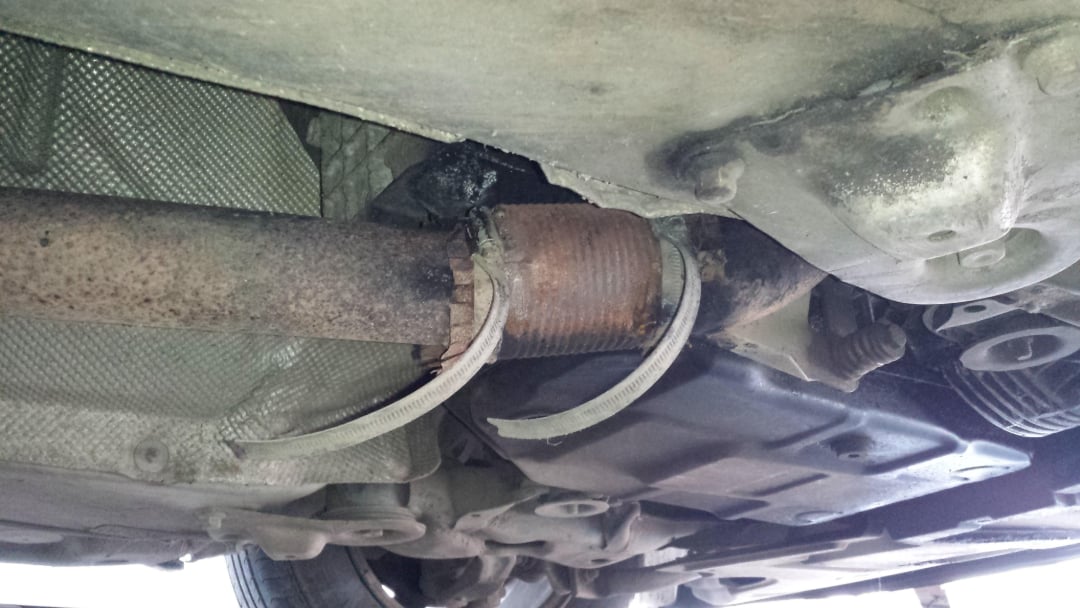A close-up view of a car’s undercarriage showing a rusty exhaust pipe with a broken flex joint and hanging metal strands.