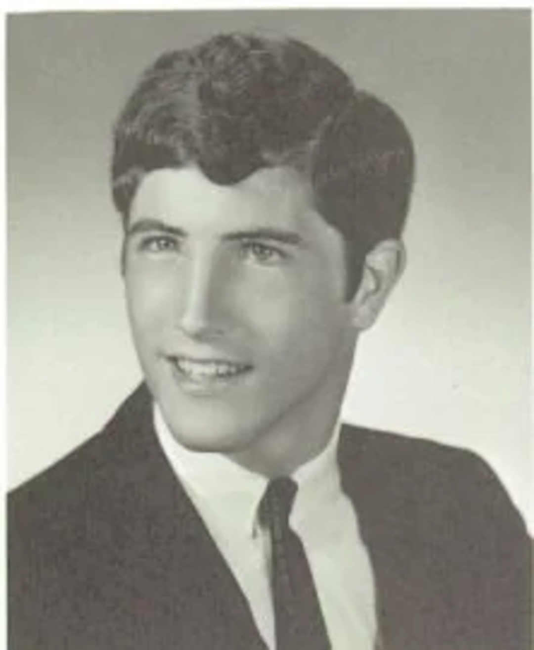 A black-and-white portrait of a young man with dark, wavy hair, wearing a suit and tie, smiling and looking slightly to the side against a plain background.