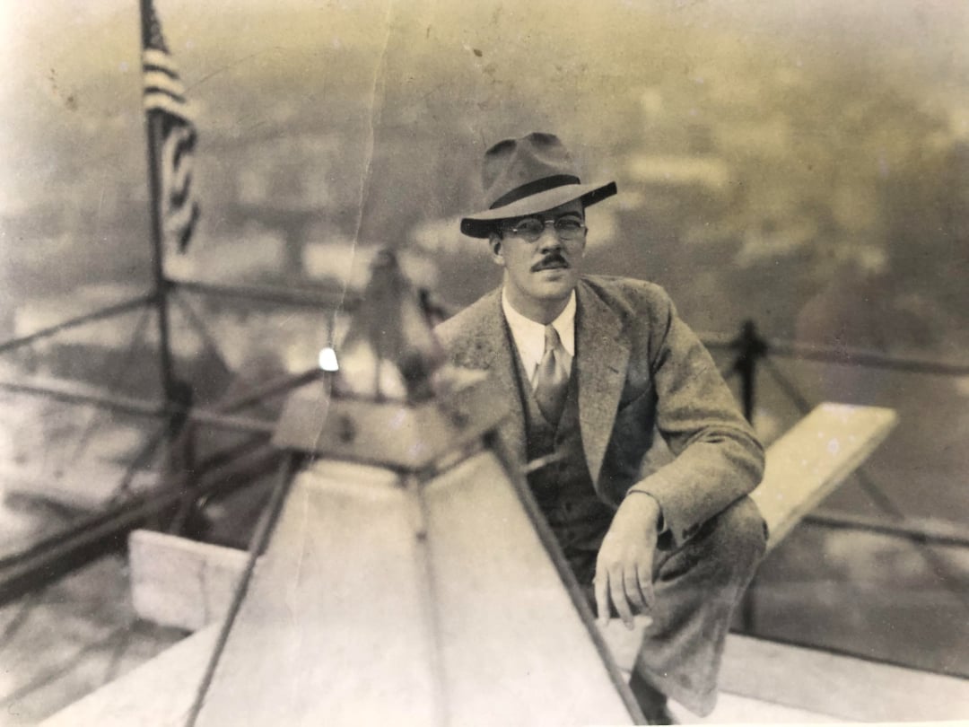 A man in a suit and hat sits on a rooftop next to a metal structure, with an American flag and a cityscape blurred in the background. The image appears vintage or from the early 20th century.