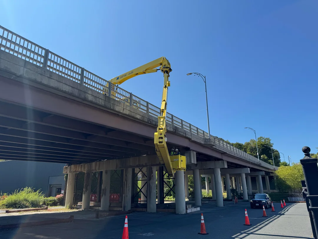 A yellow cherry picker truck with an extended arm reaches up to workers on a bridge, while orange traffic cones block off the road below on a sunny day. A car is stopped near the cones.
