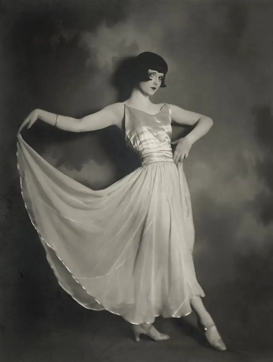 A woman in a 1920s-style, sleeveless, flowing dress poses gracefully, holding out one side of her skirt. She has a short bob haircut, dark lipstick, and stands against a softly blurred studio backdrop.