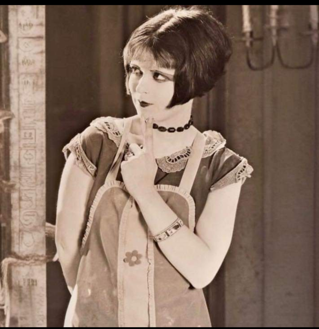 A woman with a short bob haircut, wearing a choker necklace, short-sleeved dress, and an apron with a flower design, stands indoors with one finger raised to her lips in a thoughtful pose.