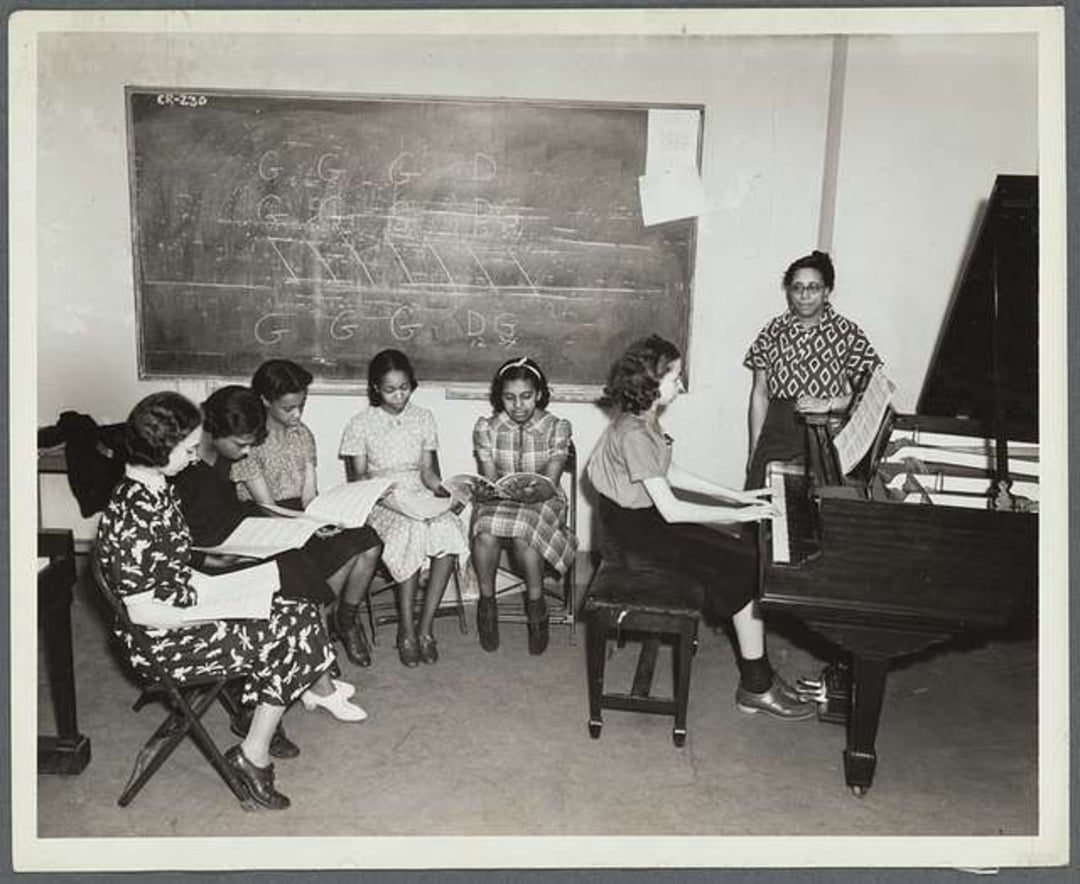 Six girls sit and sing from sheet music while another girl plays the piano; a seventh girl stands by the blackboard behind them, which shows music notes and diagrams.