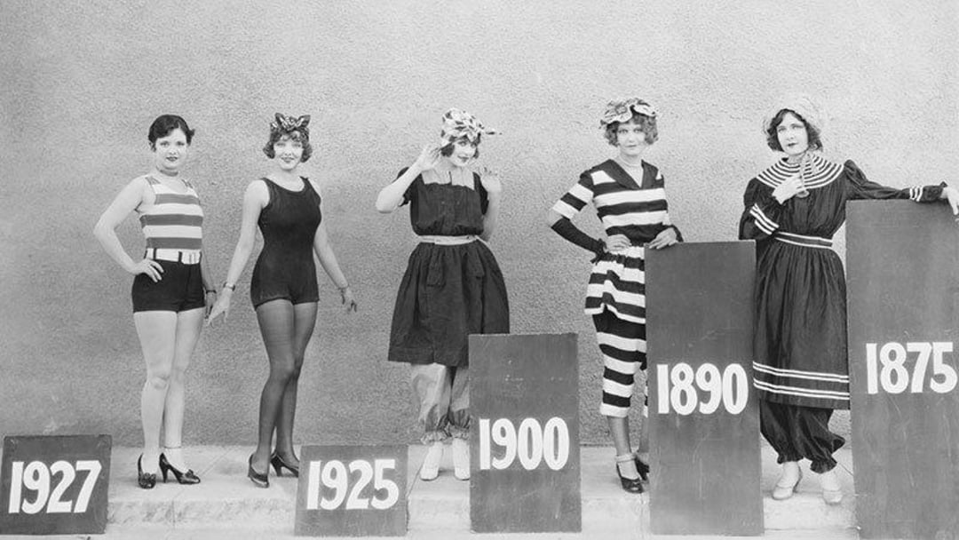 Five women model swimwear styles from 1927, 1925, 1900, 1890, and 1875, standing behind signs with those years, showing the evolution of swimsuit fashion.