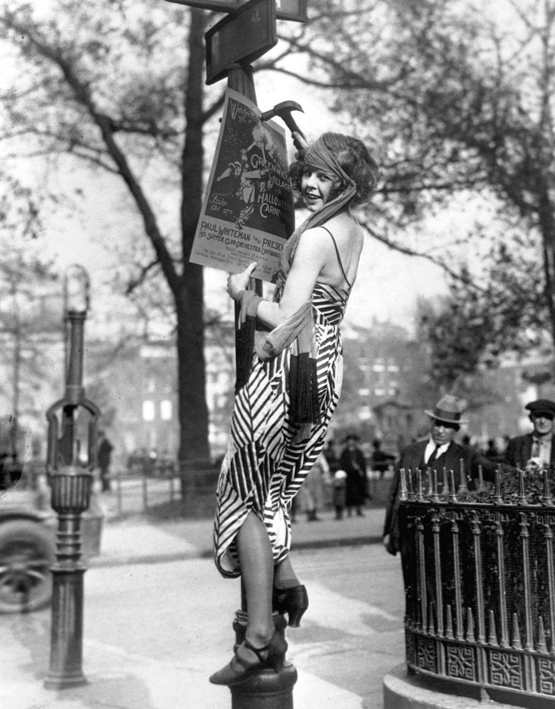 A woman in a striped dress stands on a street pole, pinning a poster to a sign in a city park. She smiles at the camera as people in early 20th-century clothing watch from the background. Trees and buildings are visible.