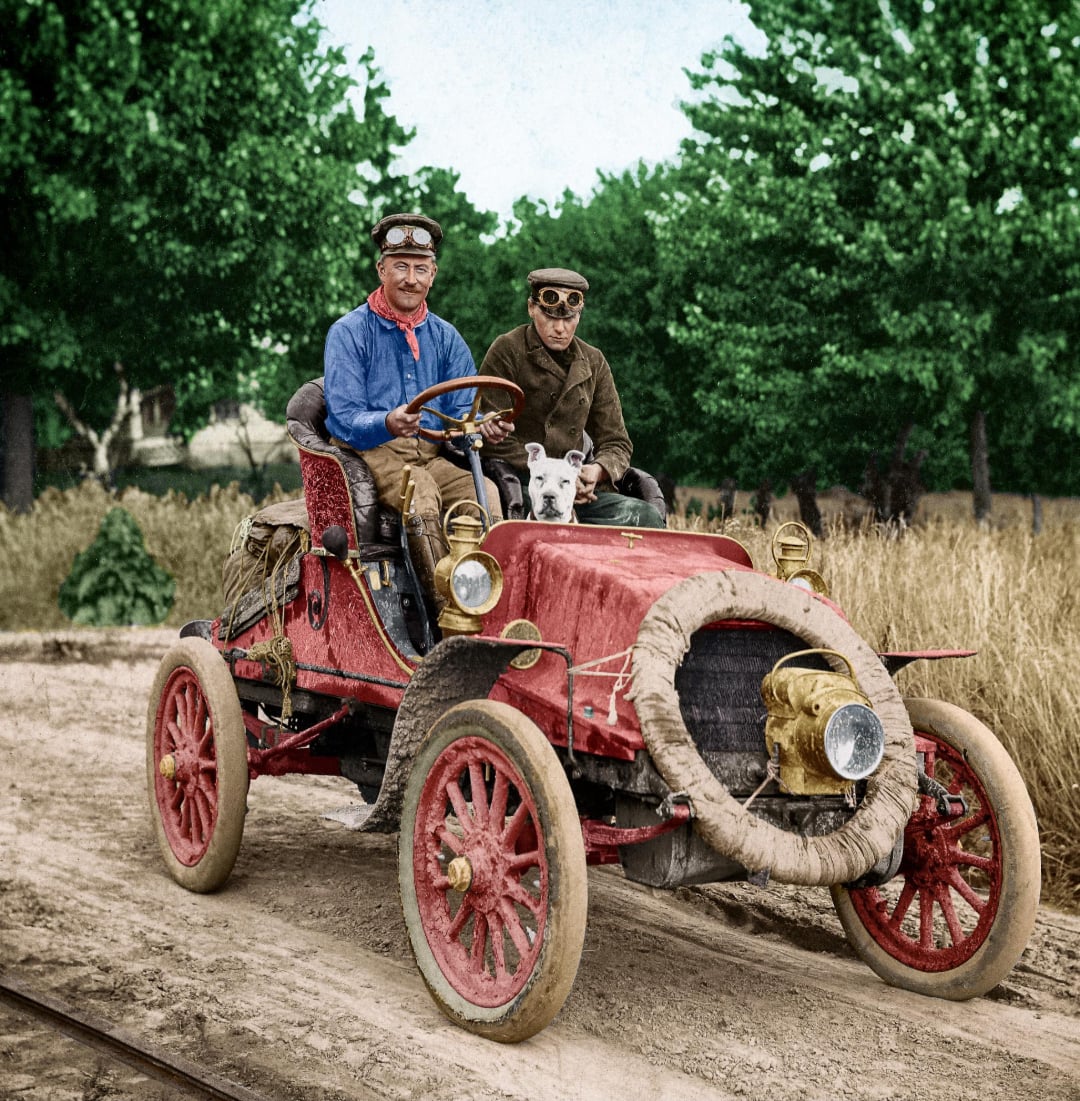 Two men wearing vintage driving goggles and hats ride an old-fashioned red car with large wheels on a dirt road, a small white dog sitting between them, with trees and tall grass in the background.