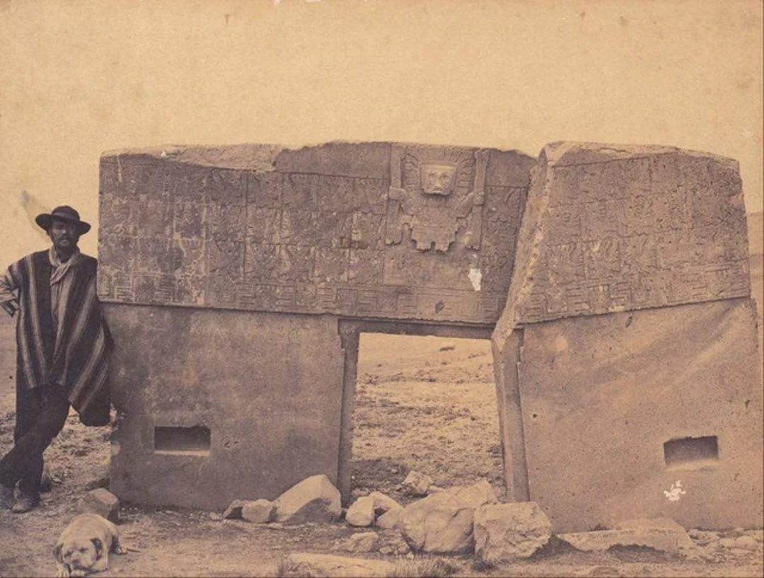 A sepia-toned photo shows a man in traditional attire and hat standing beside a large, ancient stone gateway with intricate carvings; a dog lies on the ground nearby. The structure appears weathered and is surrounded by rocks.