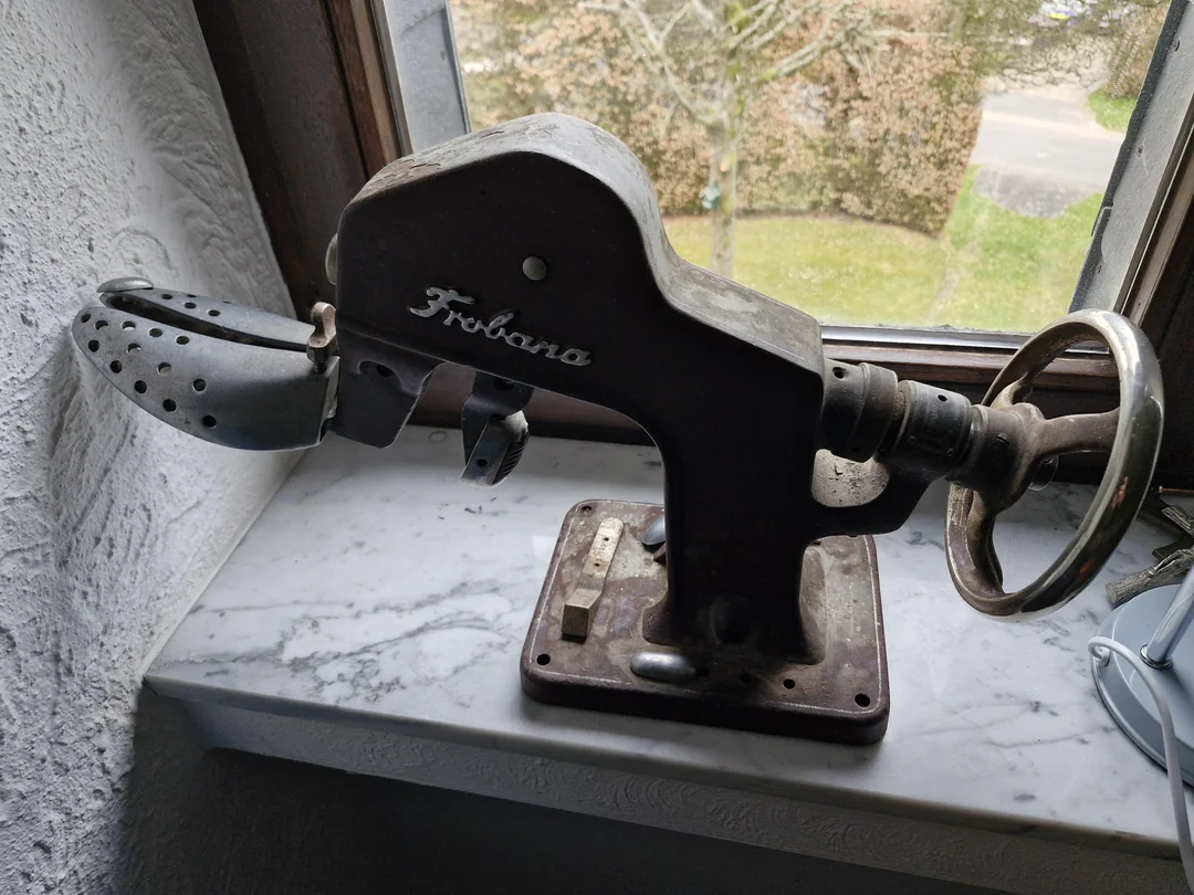 A vintage metal shoe stretcher labeled "Frubana" sits on a windowsill. The device has a rotating handle and a perforated toe form, designed to stretch and shape shoes. Natural light comes in from the window behind it.