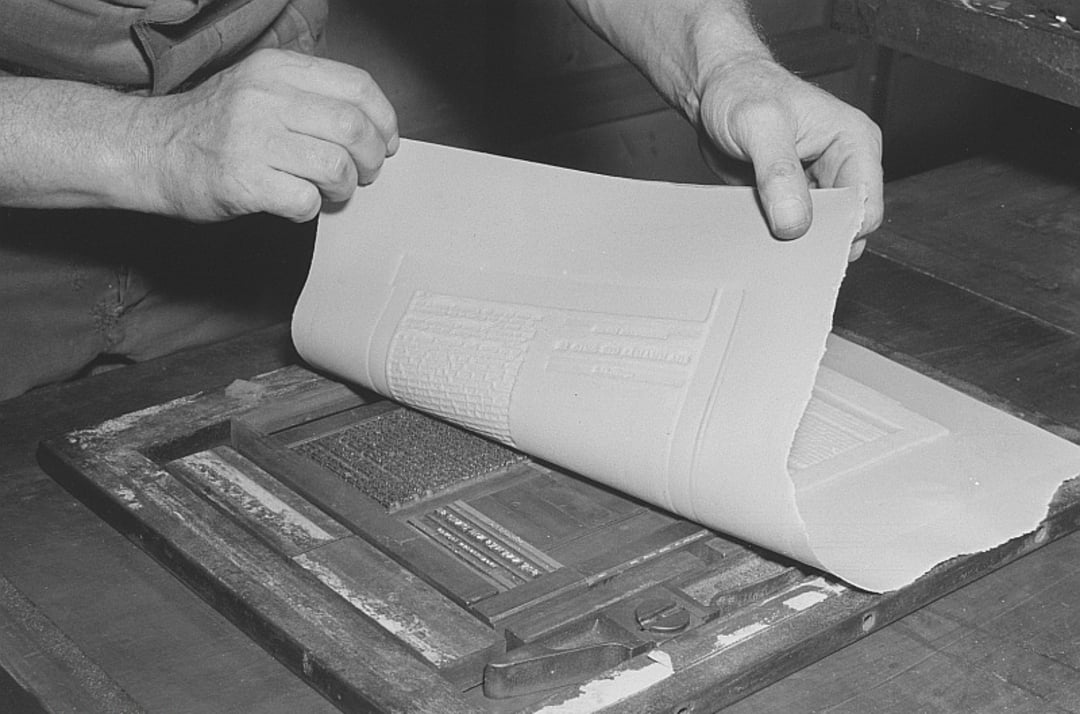 A person lifts a sheet of paper from a printing press plate, showing the process of creating prints from raised text and images on an old-fashioned press.