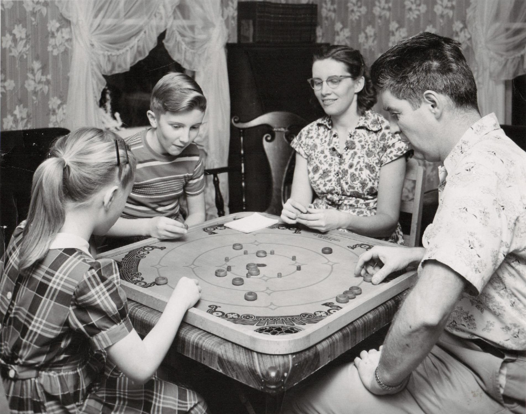 A black and white photo of a family of four playing a board game together at a square table in a living room with floral wallpaper and curtains. The mood appears focused and engaged.