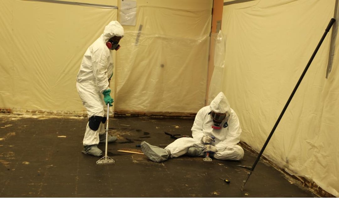 Two people in white hazmat suits and respirators remove flooring in a room with plastic-covered walls; one is standing and scraping the floor, while the other sits and works on the ground.
