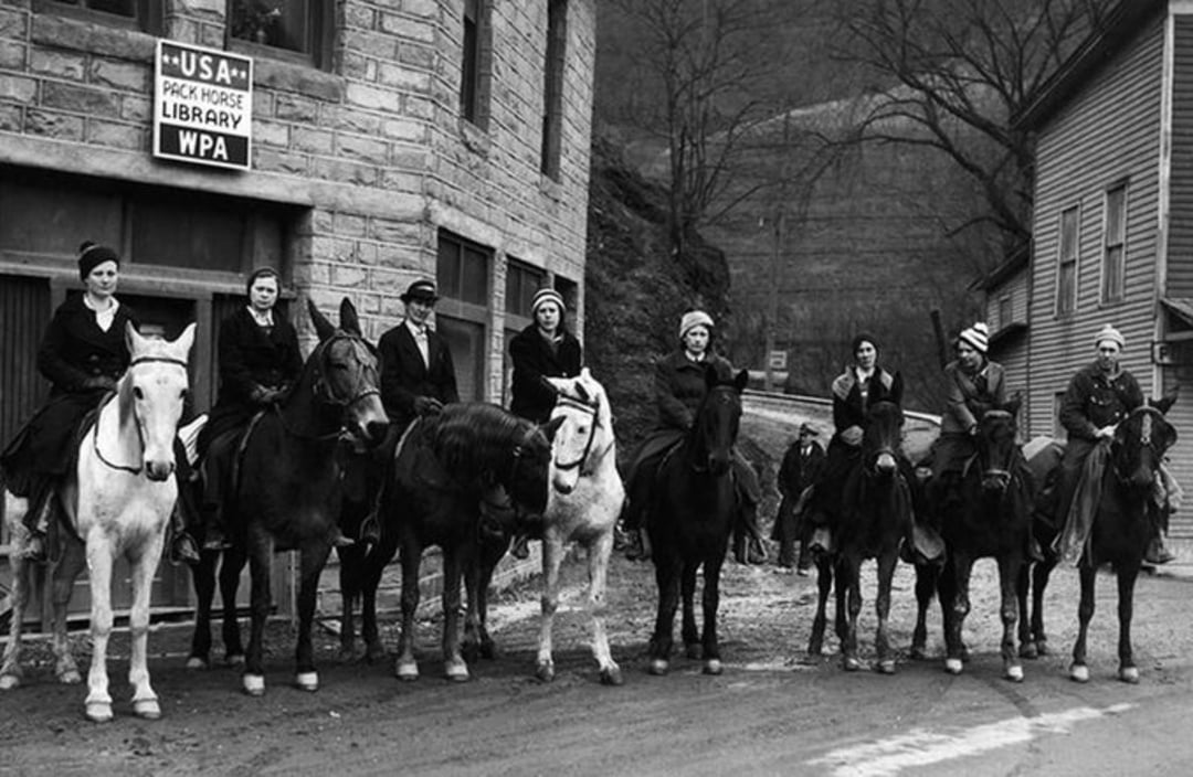 A group of eight people on horseback lines up on a street in front of a stone building with a sign reading "USA Pack Horse Library WPA" in a rural, hilly area with bare trees.