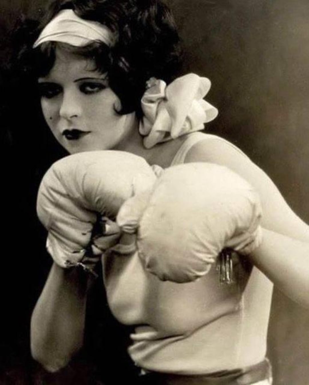 A woman with dark hair in vintage attire and large bows on her shoulders poses with oversized boxing gloves, looking determined. The image is in black and white, giving it an old-fashioned, classic feel.