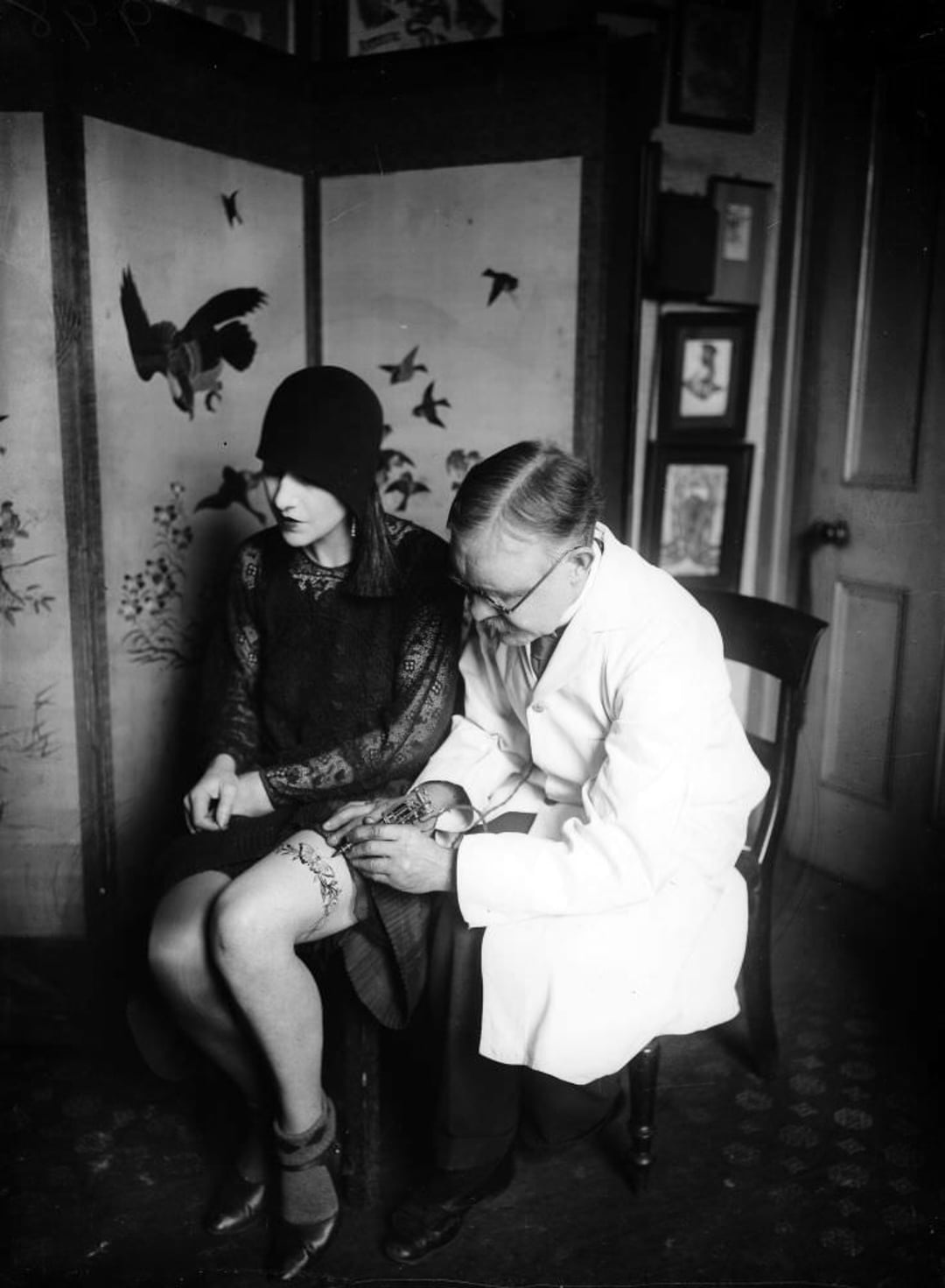 A woman in 1920s attire sits while a man in a white coat tattoos her knee. They are indoors, next to a decorative folding screen with bird illustrations. The woman looks down at the tattoo being applied.