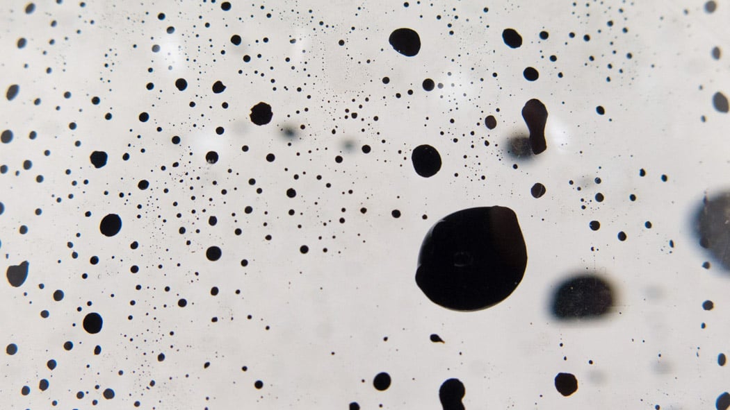 Black ink droplets of various sizes scattered randomly on a white background, creating an abstract, speckled pattern.