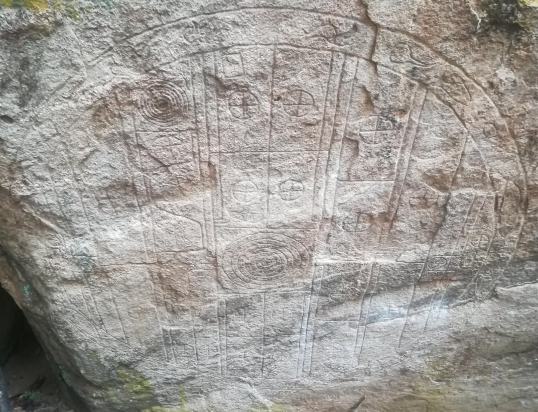 Weathered stone slab with intricate carvings, including spirals, circles, and vertical lines, likely representing ancient or prehistoric rock art. The surface appears rough and partially eroded by time.