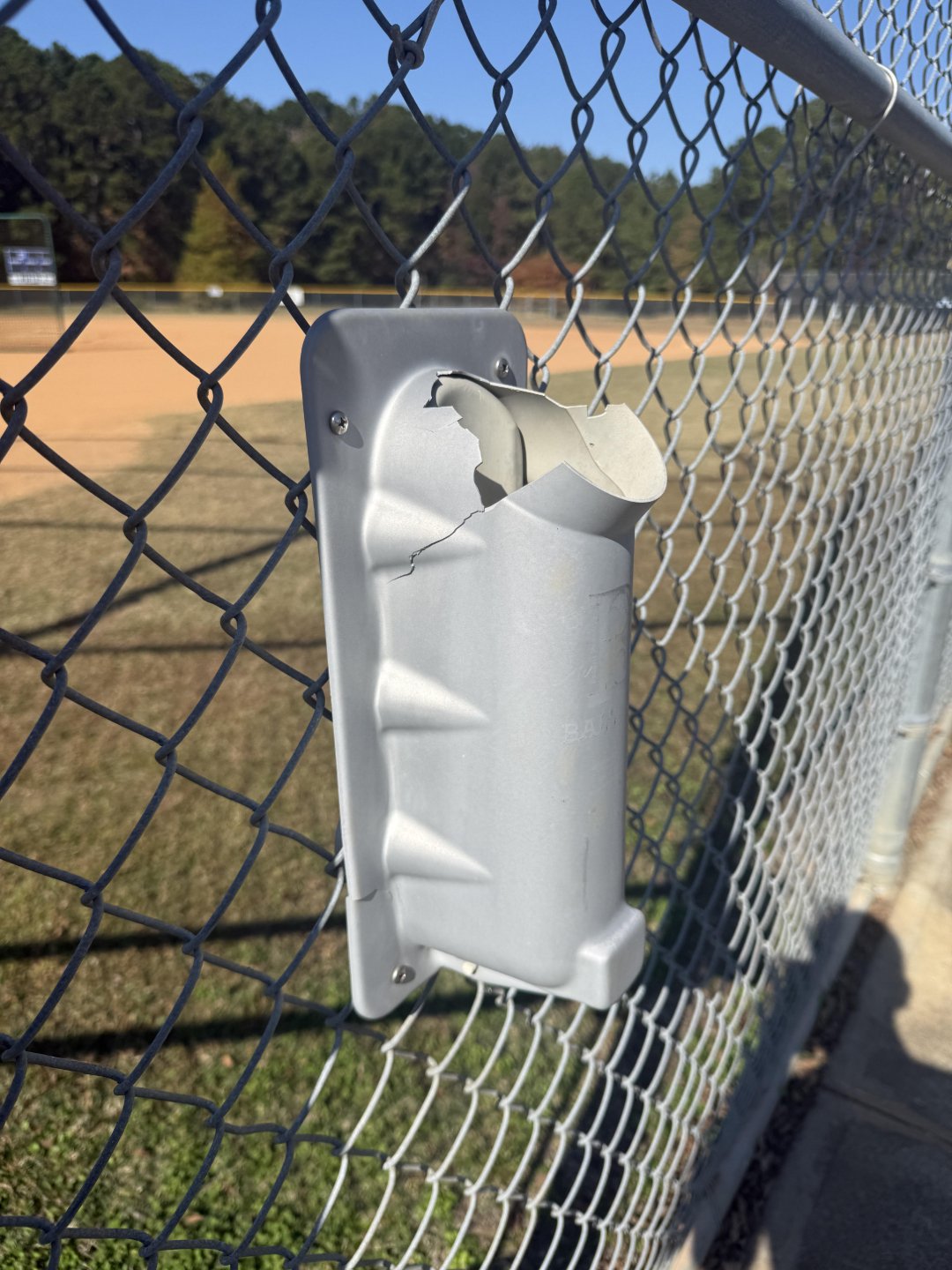 A broken silver baseball bat holder attached to a chain-link fence at a baseball field, with trees and a grassy area visible in the background.