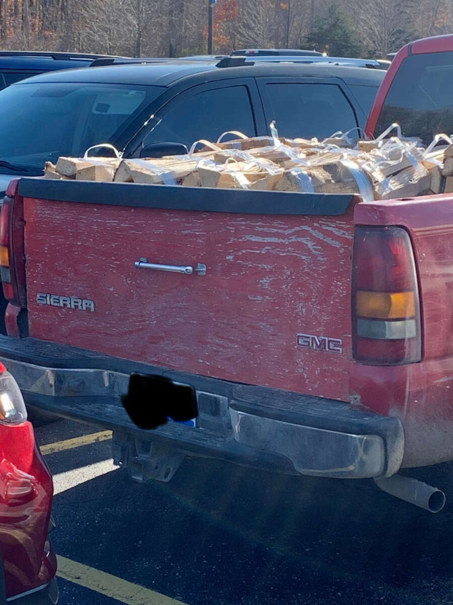 A red GMC Sierra pickup truck with a worn tailgate is parked in a lot, its bed loaded with several bundled stacks of firewood. The license plate is partially obscured.