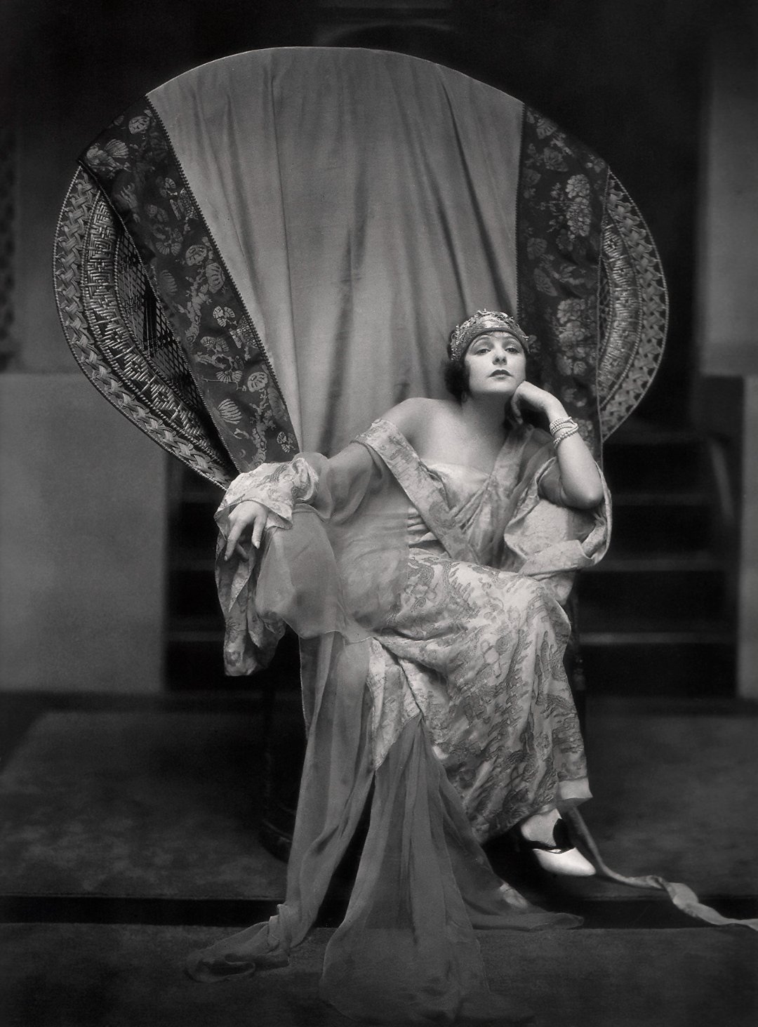 A woman in an ornate gown and headpiece sits on a large, round-backed chair draped with patterned fabric, gazing thoughtfully to the side with one arm resting on the chair and the other on her face.
