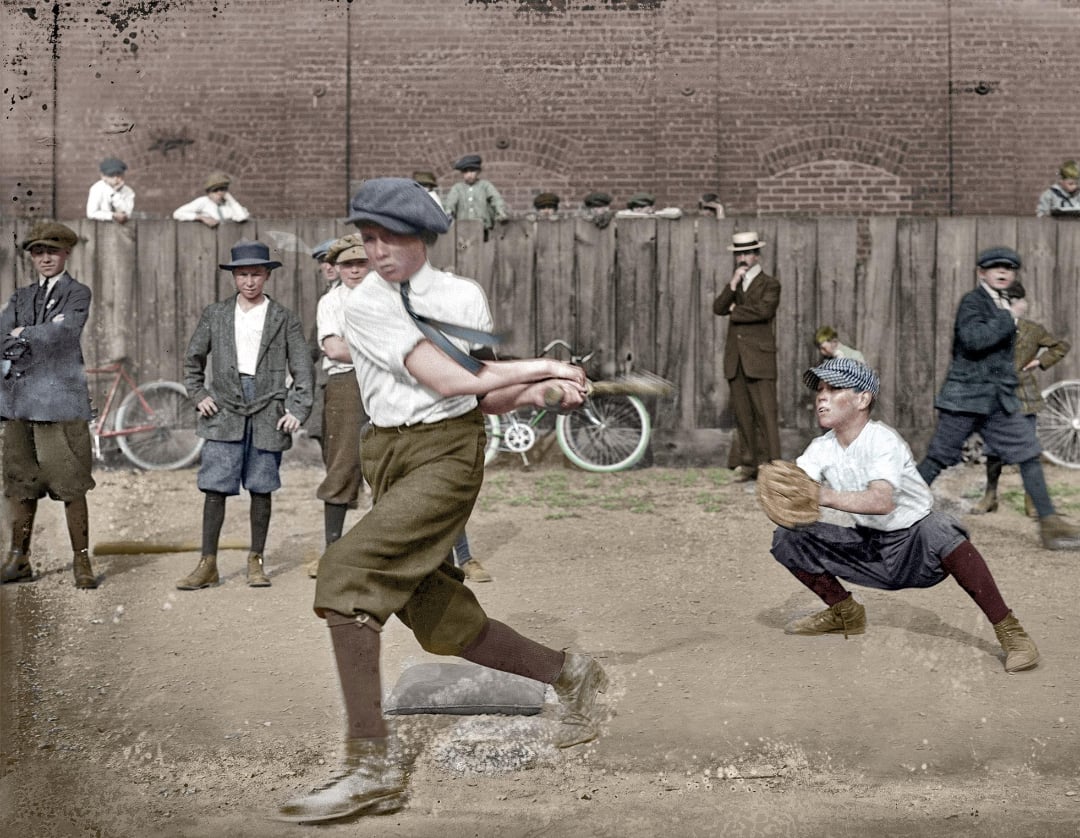 A group of boys in early 20th-century clothing play baseball on a dirt lot, with one swinging a bat and another catching. Bystanders watch near a wooden fence, and bicycles lean against the wall in the background.