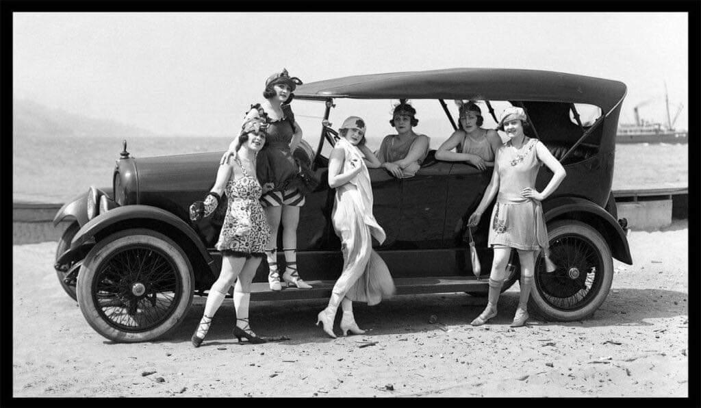 Five women in 1920s flapper dresses pose by a vintage car on a beach. Some are sitting on the car, while others stand or lean against it. A body of water and a ship are visible in the background.