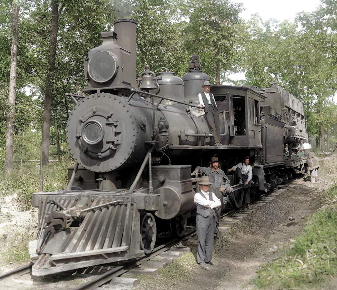 A vintage steam locomotive on railroad tracks in a wooded area, with four men in early 20th-century clothing standing beside and on the train. The scene is in daylight and surrounded by trees.