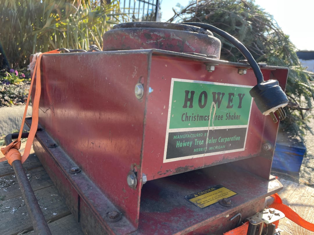 A red HOWEY Christmas Tree Shaker machine with a green and white label sits outdoors. An attached power plug rests on top, and a pile of tree branches is visible in the background.