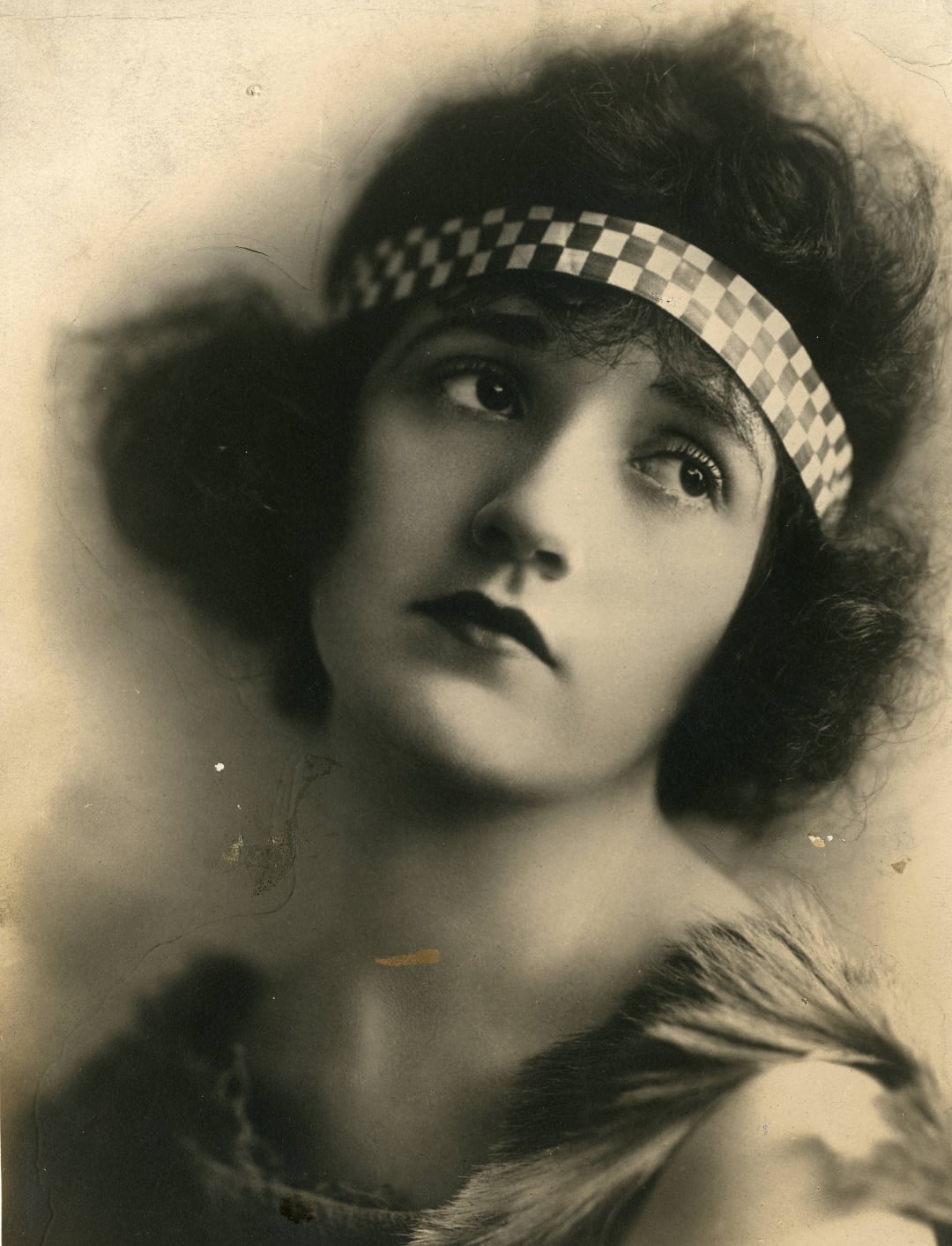 Black and white portrait of a woman with short wavy hair, wearing a checkered headband and a fur-trimmed garment, gazing upward with a thoughtful expression.