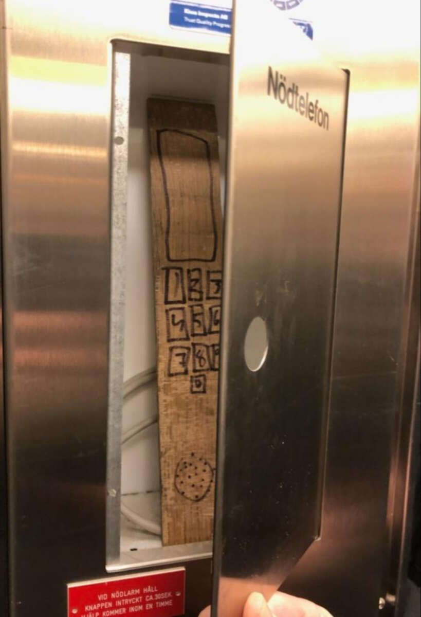 A metal emergency phone box labeled "Nödtelefon" is open, revealing a wooden plank with a phone keypad and speaker crudely drawn on it with marker instead of an actual telephone.