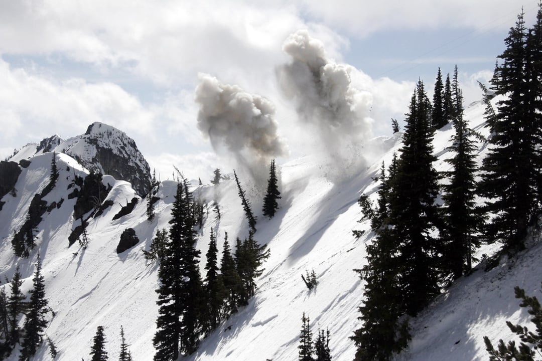 A snowy mountain slope with tall pine trees and clouds of smoke rising, suggesting an avalanche or controlled explosion, under a partly cloudy sky.