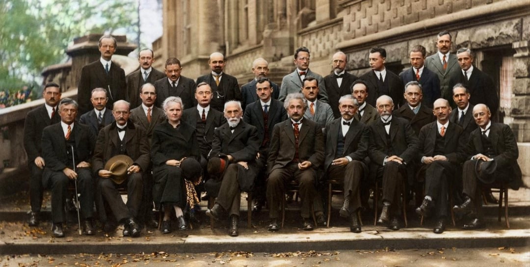 A group of men and one woman in formal attire pose for a historic black-and-white photo, seated and standing in rows outside a stone building, likely at a scientific or academic gathering.