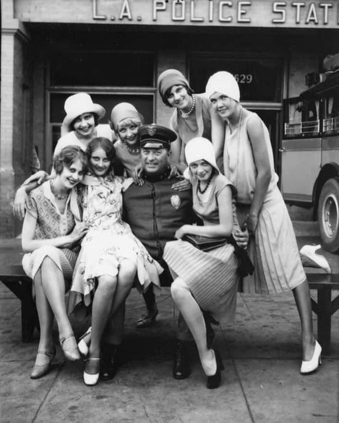 Seven smiling women in 1920s flapper dresses and hats pose around a seated police officer outside an L.A. Police Station. Everyone appears happy and relaxed, with vintage cars and the station building in the background.