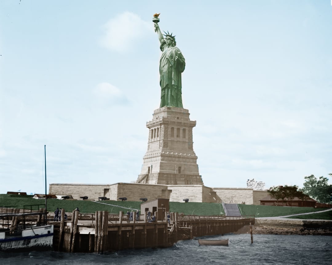 The Statue of Liberty stands tall on its pedestal, with a torch raised high, seen from the waterfront. A dock, people, and small boats are visible in the foreground under a partly cloudy sky.