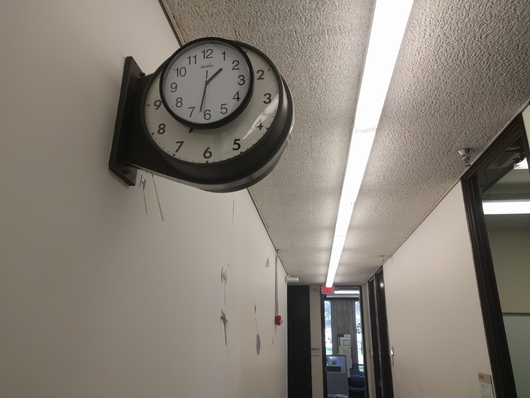 A black-framed wall clock showing 9:22 is mounted in a white hallway with a textured ceiling and fluorescent lights. There are doors and windows along the corridor and some marks on the wall below the clock.
