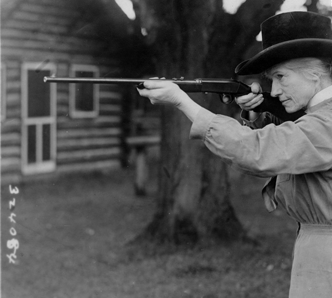 An older woman wearing a hat and long-sleeved dress aims a rifle outdoors, with trees and a wooden cabin visible in the background.