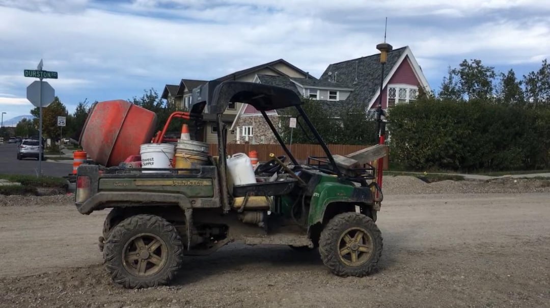 A utility vehicle with muddy tires is parked on a dirt road, carrying buckets, construction tools, and an orange concrete mixer in the back. Residential houses and trees are visible in the background.