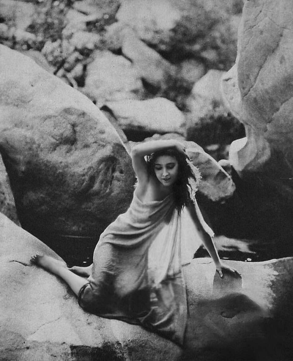 A woman in a flowing dress sits on large rocks near water, resting one hand on her head and the other on a rock. The image is black and white, with soft, natural light and an ethereal, vintage atmosphere.